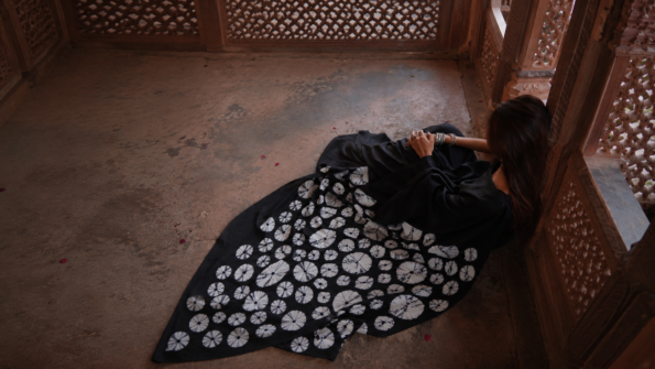 Woman in elegant black printed saree sitting in a heritage-style corridor with artistic monochrome fabric spread and warm natural lighting