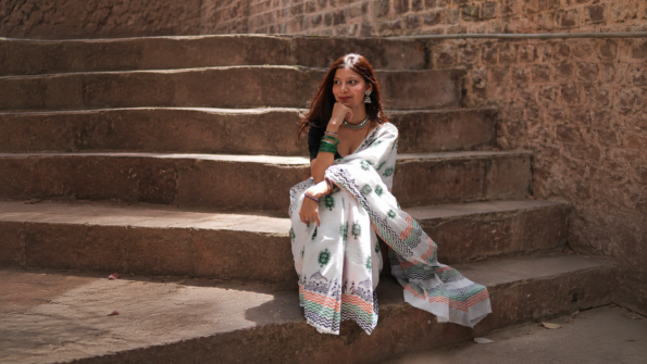 Woman wearing a white printed saree sitting on rustic stone steps in warm natural sunlight with traditional silver jewelry
